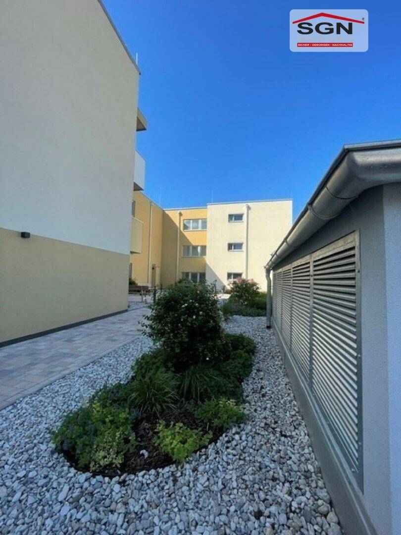 Schmaler Weg zwischen zwei Gebäuden einer Wohnanlage mit Kiesbeet und Lüftungsgittern an der Wand.