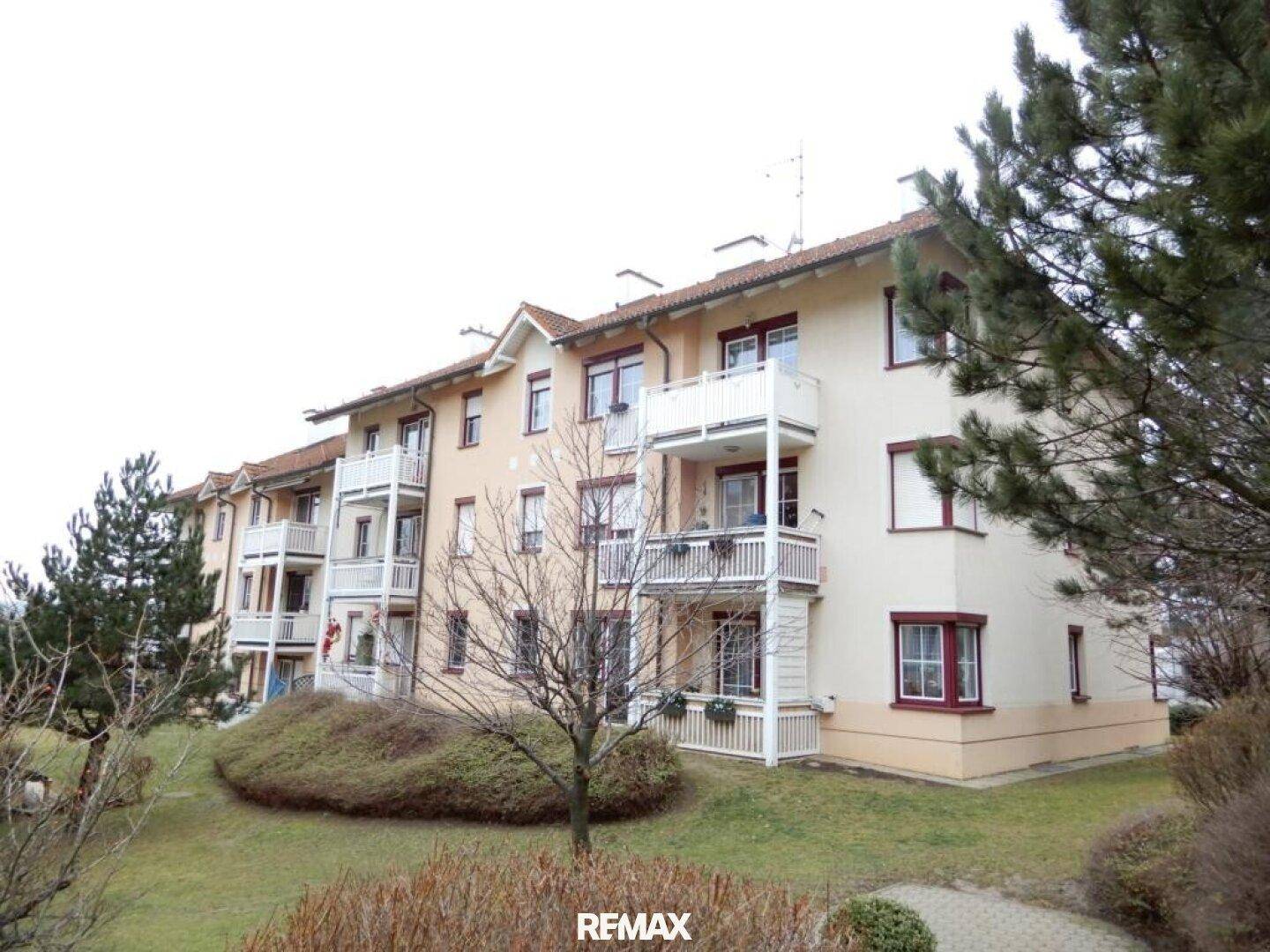 Mehrfamilienhaus mit mehreren Balkonen, gepflegter Grünanlage und Bäumen im Vordergrund.