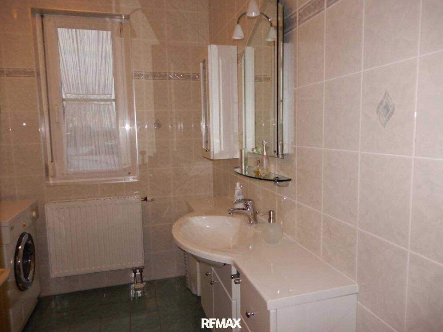 Gefliestes Badezimmer mit Waschbecken, Spiegelschrank, Fenster und Heizkörper.
