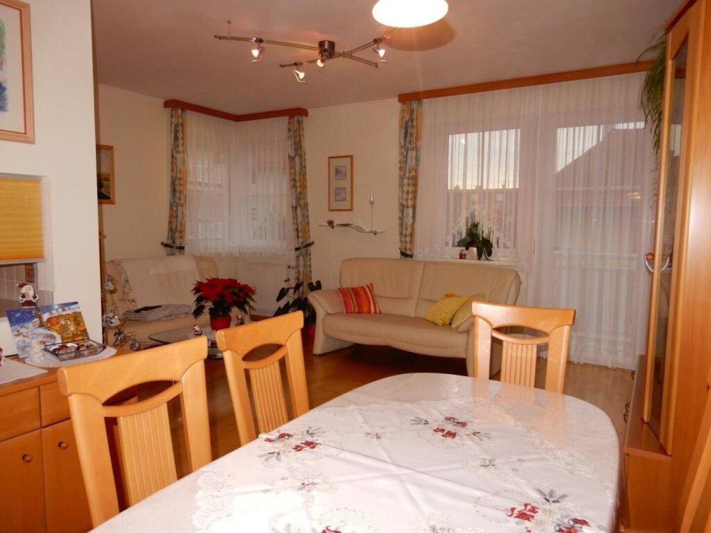 Gemütlicher Wohn- und Essbereich mit Holzboden, beiger Couch und großen Fenstern.