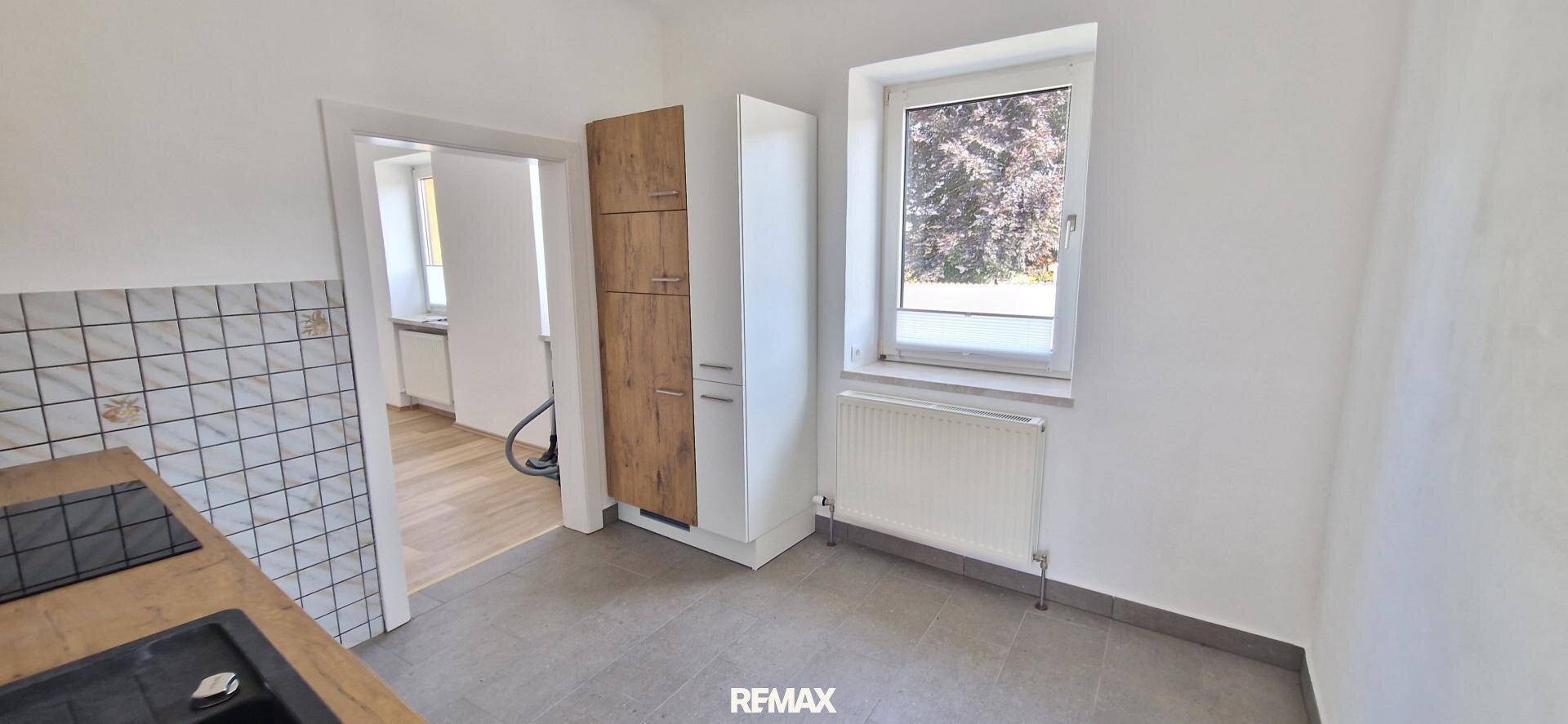Helle Küche mit Fenster, Heizkörper und Blick in einen angrenzenden Raum mit Holzboden.