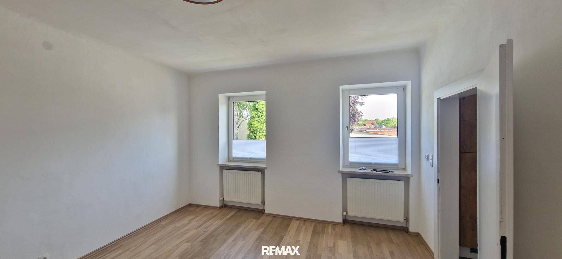 Geräumiges, leeres Zimmer mit hellem Holzboden, zwei Fenstern und viel Tageslicht.