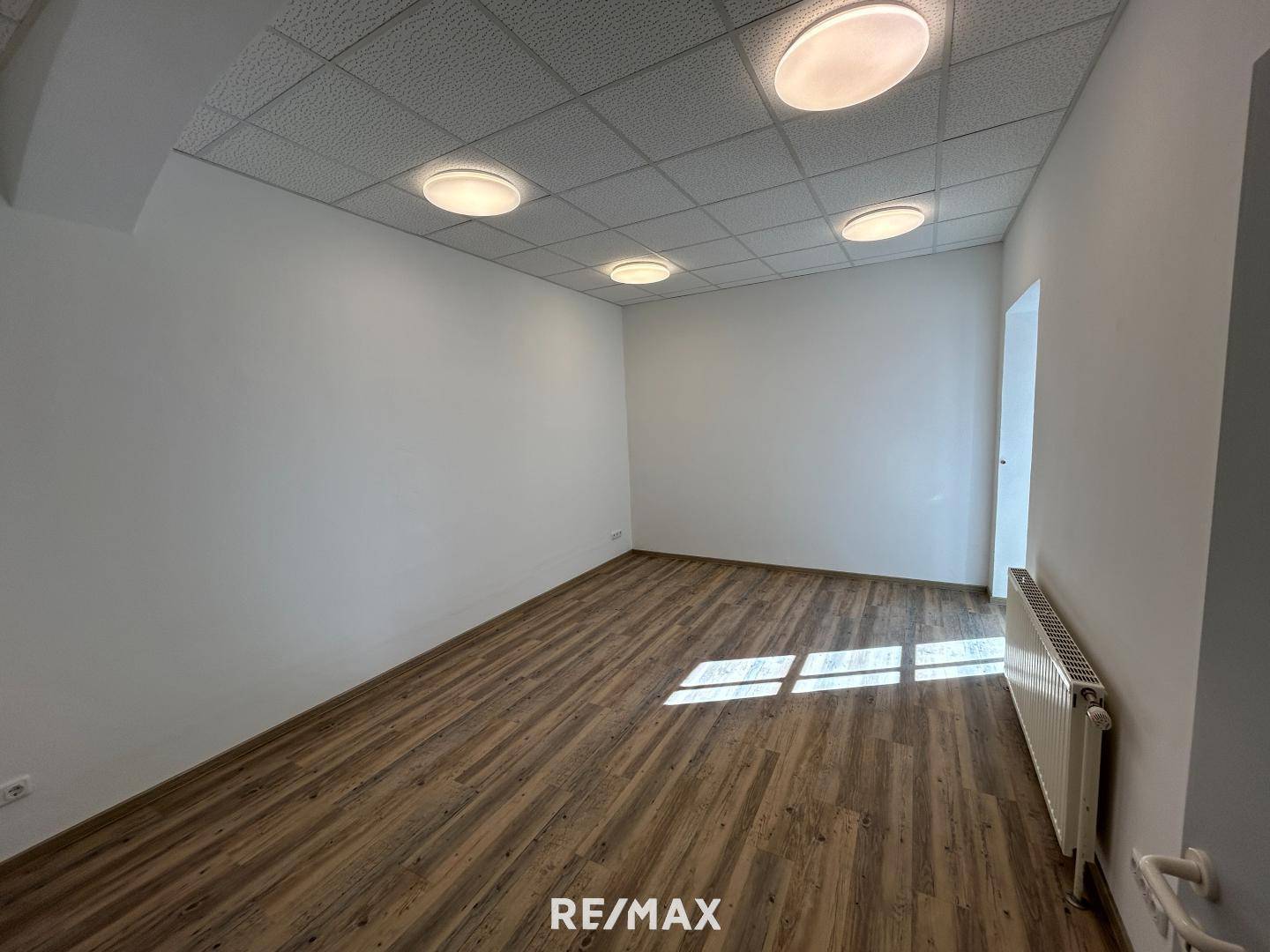 Heller Büroraum mit Laminatboden, abgehängter Decke und natürlichem Lichteinfall durch ein Fenster.