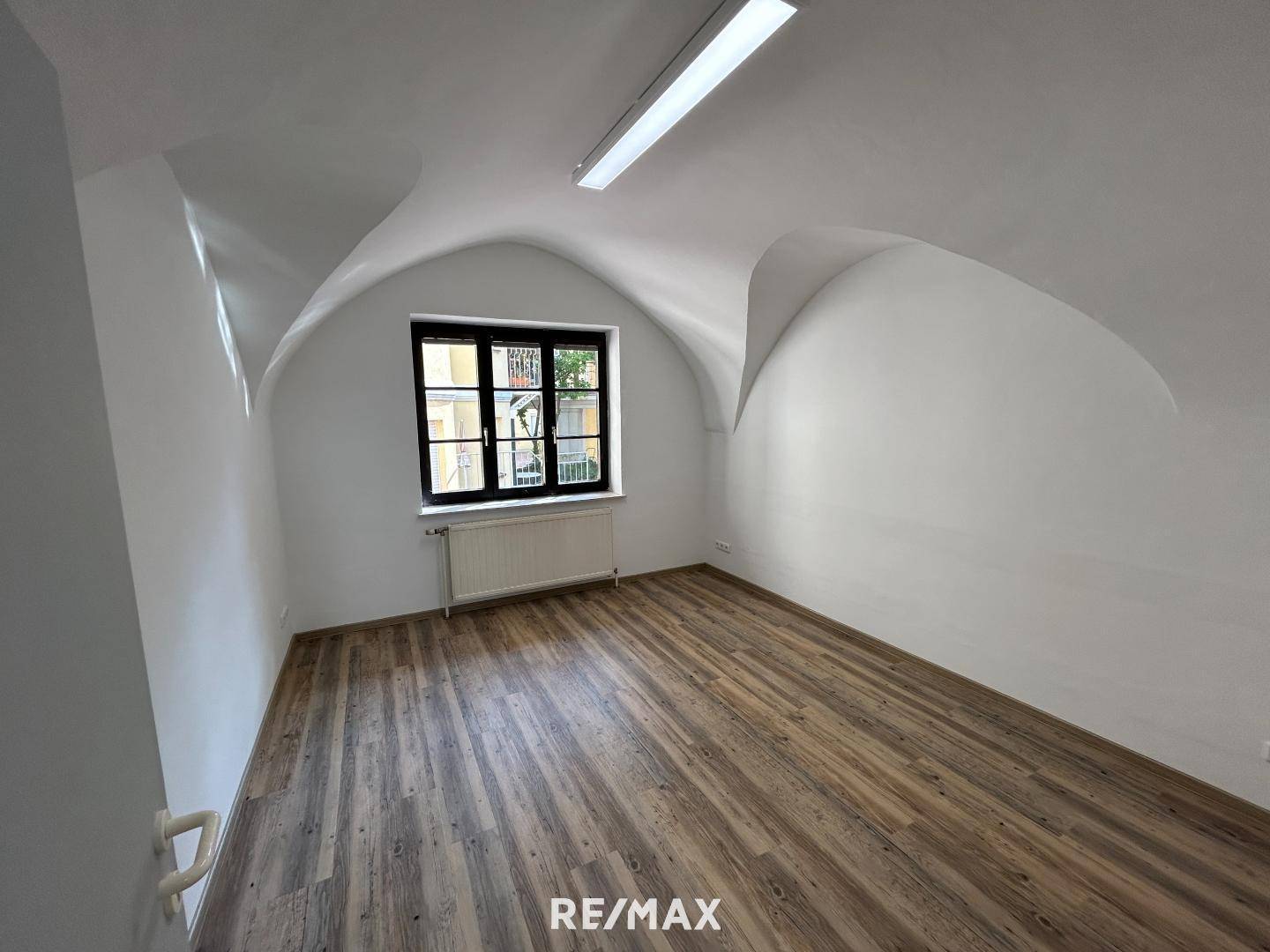Heller Büroraum mit Gewölbedecke, Laminatboden und einem Fenster mit Blick nach draußen.