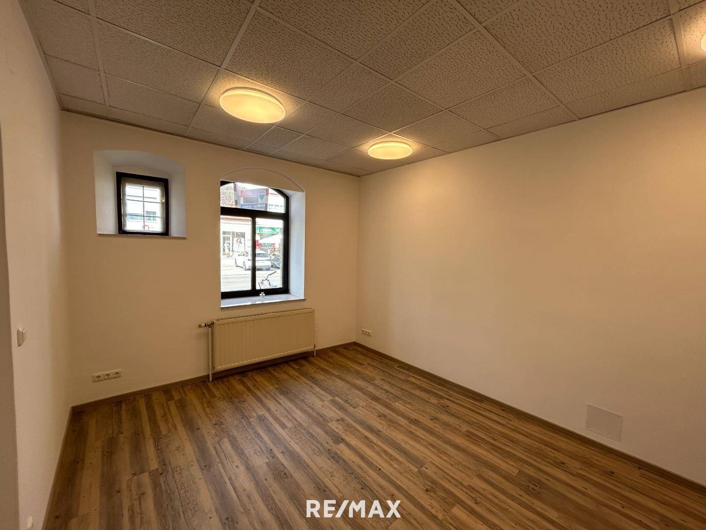 Heller Büroraum mit Laminatboden, abgehängter Decke und zwei Fenstern zur Straße.