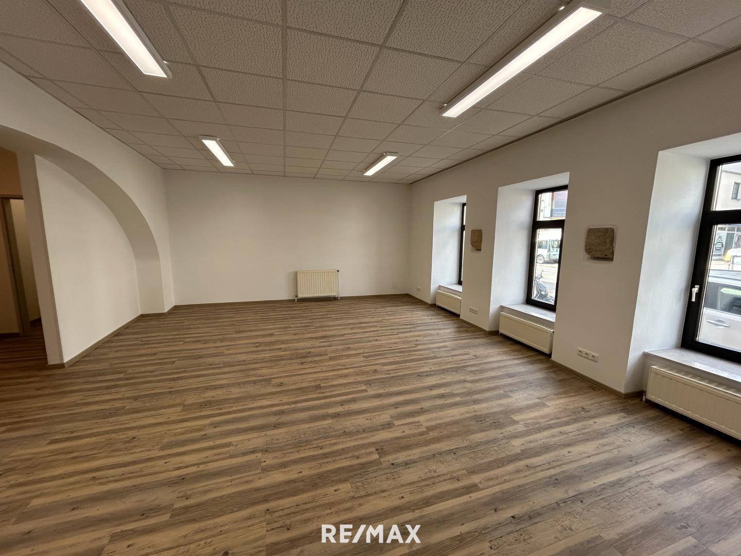 Großer, heller Raum mit Laminatboden, abgehängter Decke und mehreren Fenstern zur Straße.