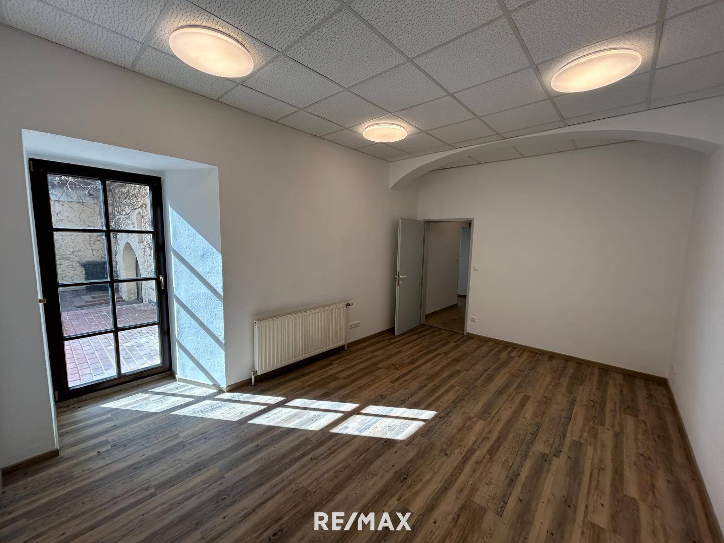 Geräumiger Büroraum mit Laminatboden, abgehängter Decke und großem Fenster zum Innenhof.