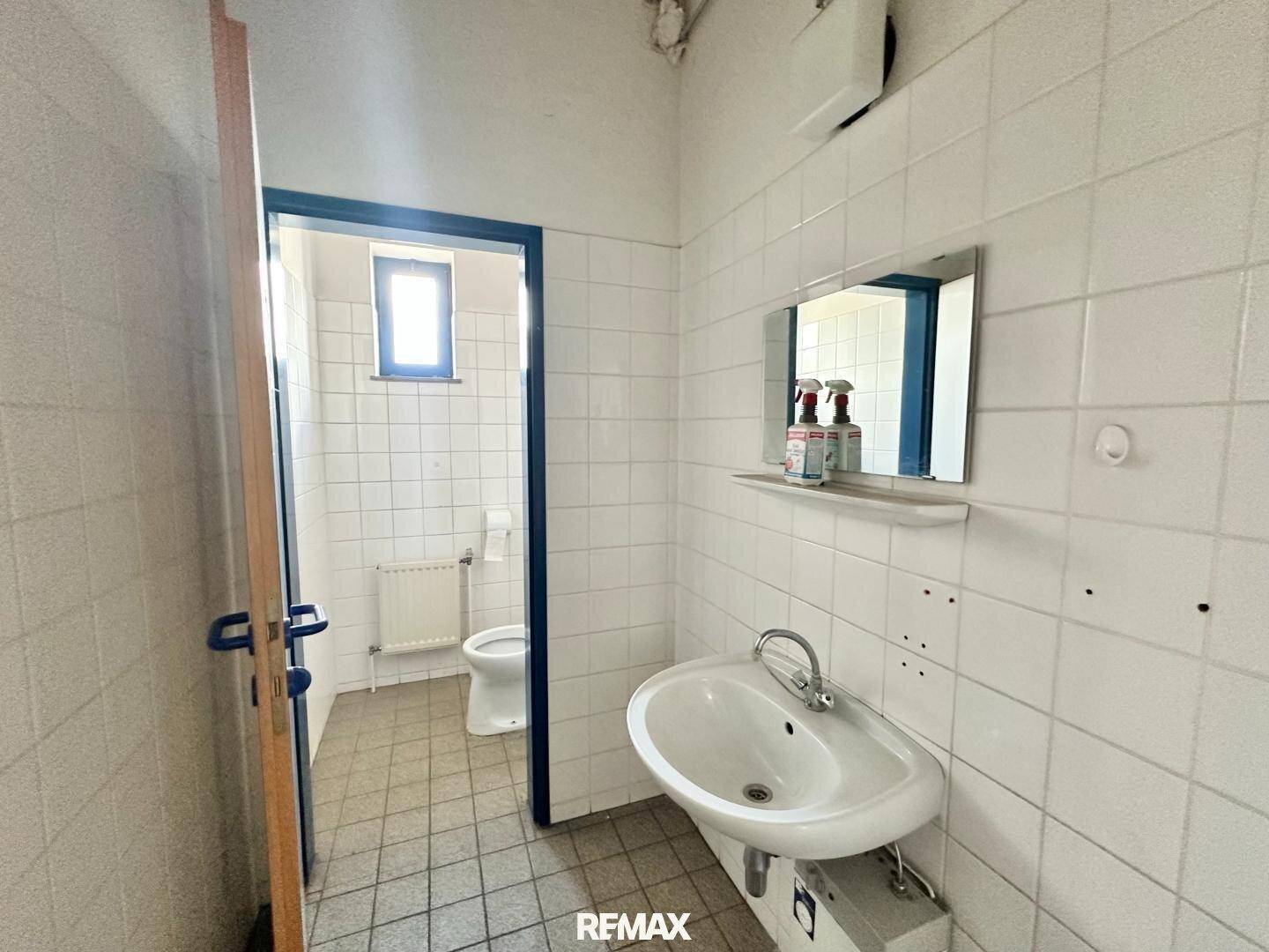 Badezimmer mit weißen Fliesen, Waschbecken und einer Toilette, sichtbar durch die offene Tür.