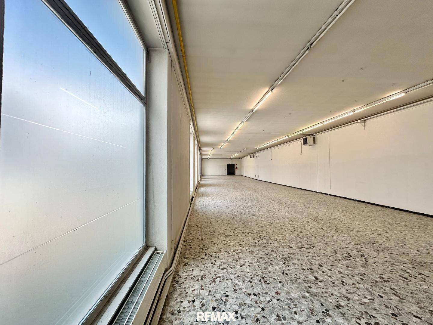 Langer, leerer Raum mit großen, undurchsichtigen Fenstern und Terrazzo-ähnlichem Bodenbelag.