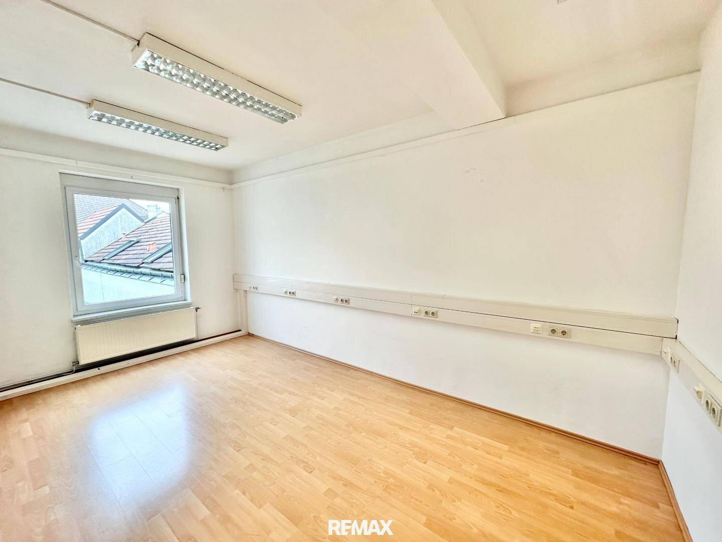 Heller Büroraum mit Holzparkettboden, weißen Wänden und Fenster mit Blick auf Dächer und Himmel.