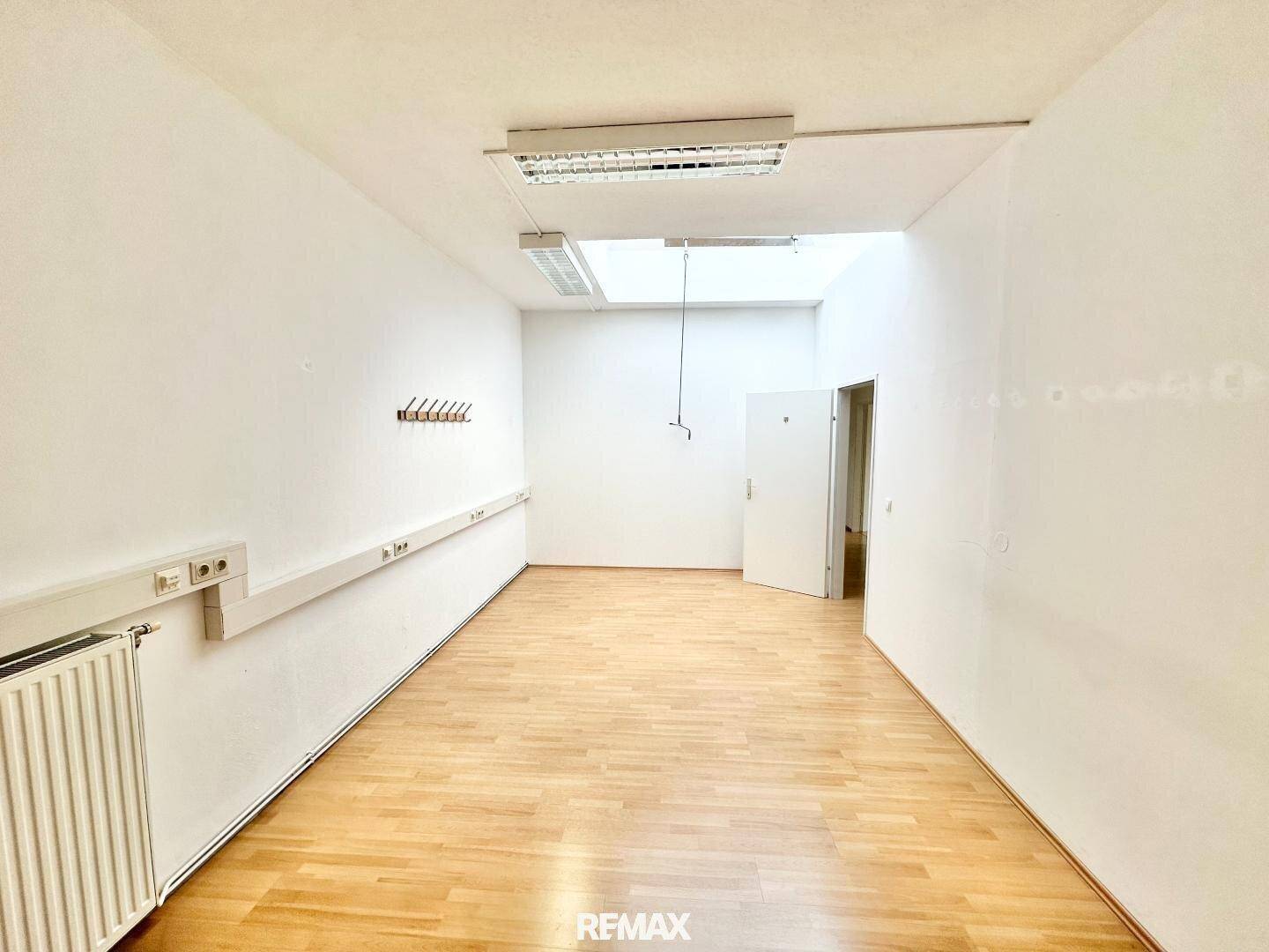 Geräumiger Büroraum mit Holzparkettboden, weißen Wänden und Garderobe, mit viel Tageslicht durch Oberlicht.