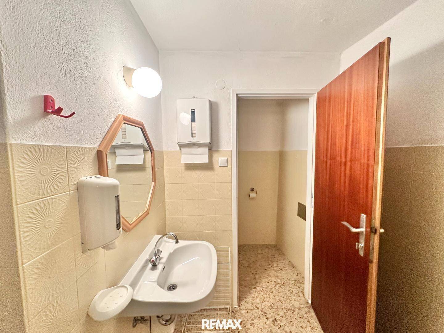 Badezimmer mit Waschbecken, Spiegel und hellen Fliesen, führt zu einer separaten Toilettenkabine.