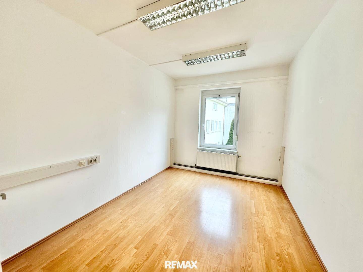 Heller Büroraum mit Holzparkettboden, weißen Wänden und Fenster mit Blick auf die Umgebung.
