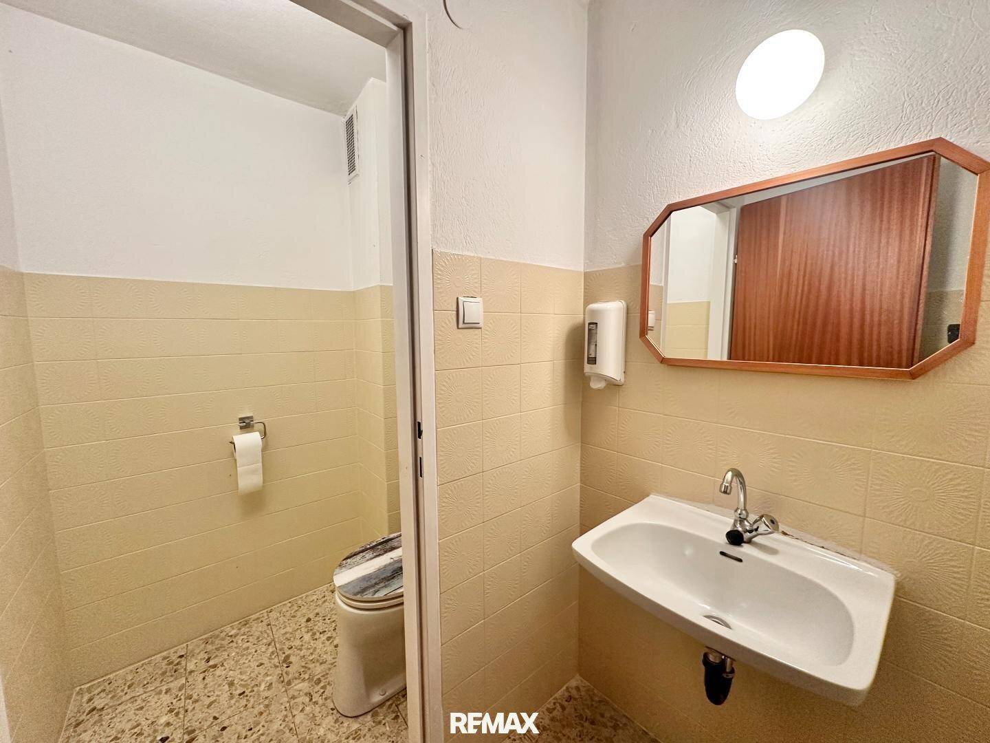 Badezimmer mit Waschbecken, Spiegel und Toilette, ausgestattet mit hellen Fliesen und Spender.