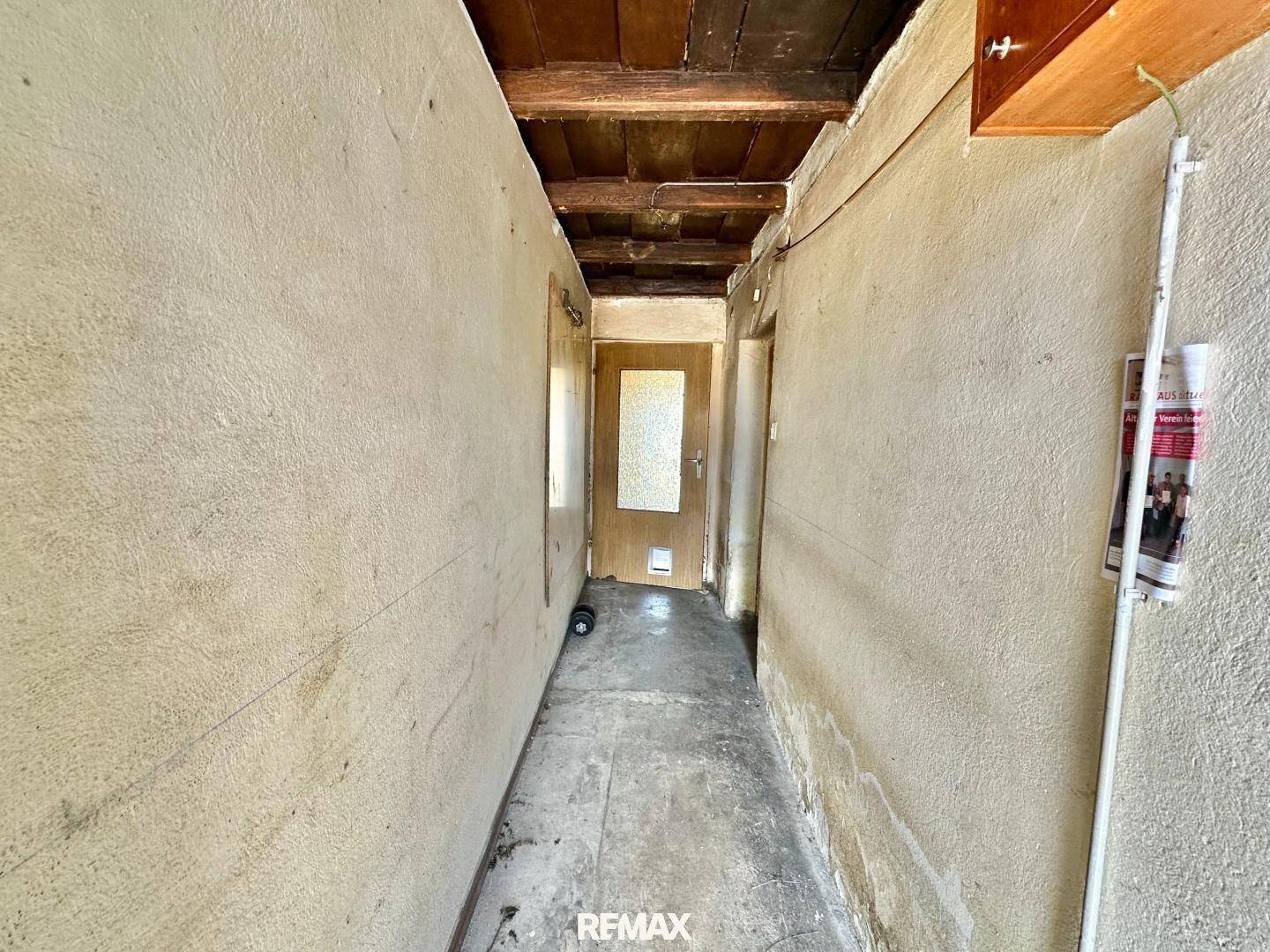 Schmaler Flur mit unverputzten Wänden, sichtbaren Holzdeckenbalken und einer einfachen Holztür am Ende.