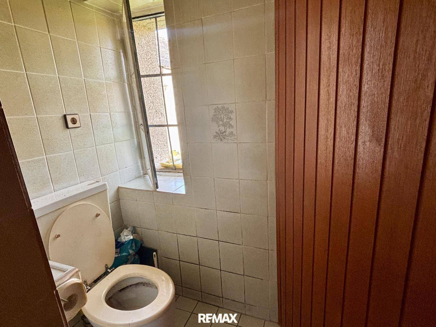 Einfache Toilette mit Fliesen an den Wänden und einem kleinen Fenster, das Renovierung benötigt.
