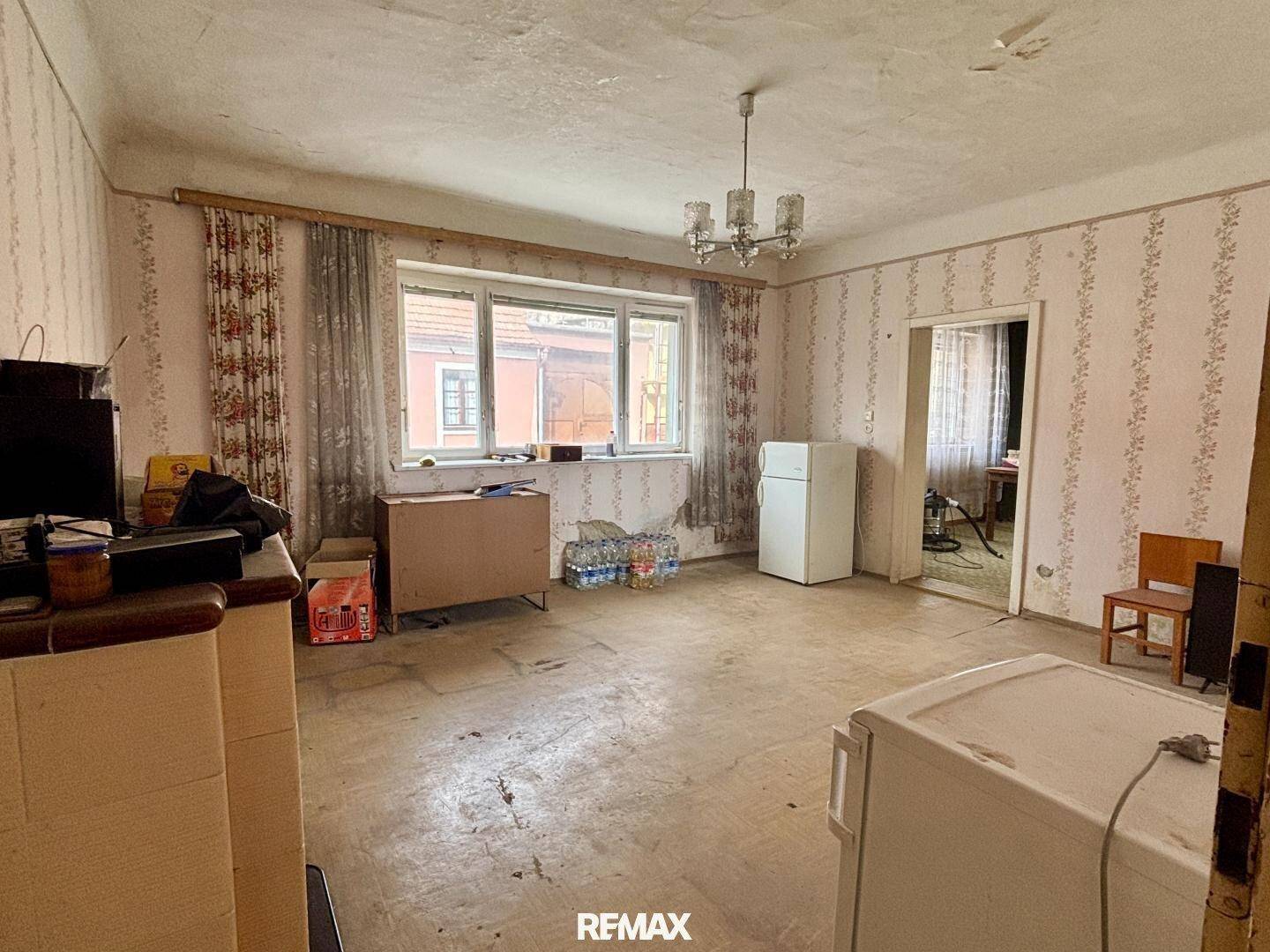 Geräumiges Wohnzimmer mit abgenutztem Boden, Tapeten und einem Fenster zur Straße, das Renovierung benötigt.