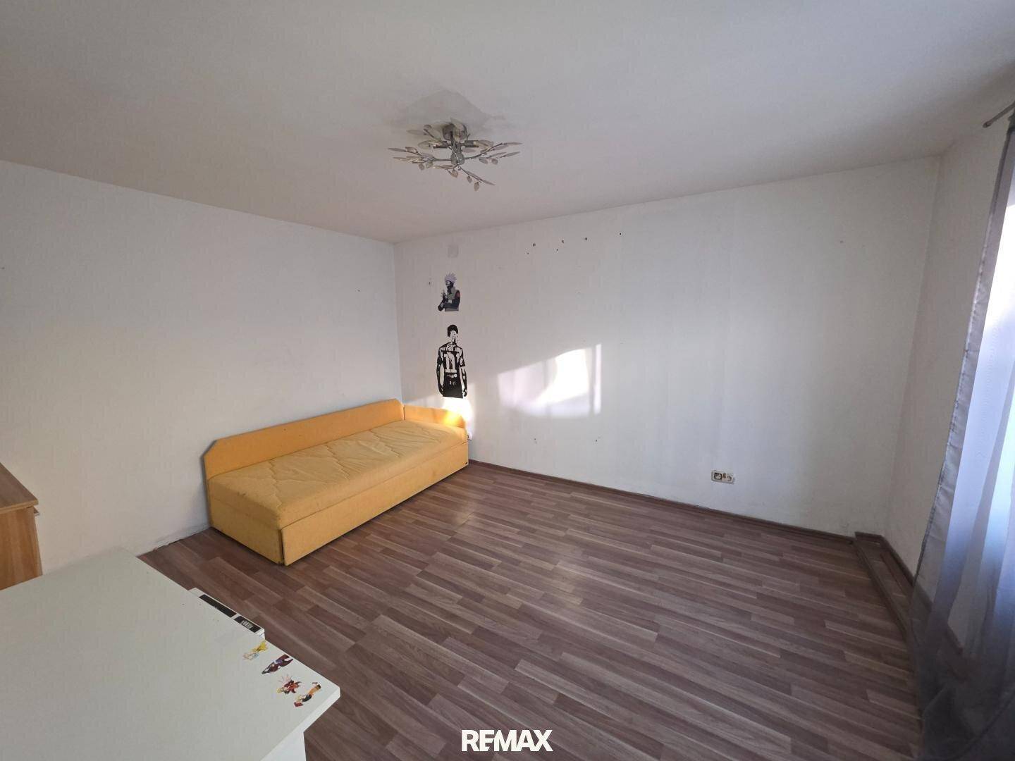 Schlafzimmer mit dunklem Laminatboden und einem gelben Schlafsofa unter einem Fenster.