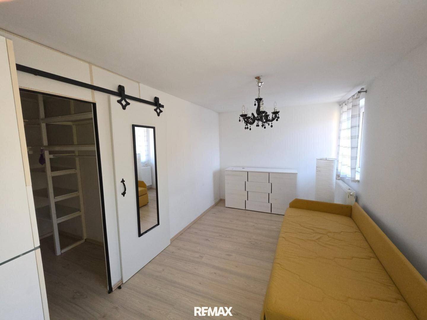 Schlafzimmer mit hellem Laminatboden, gelbem Sofa und Schiebetür zu einem begehbaren Kleiderschrank.
