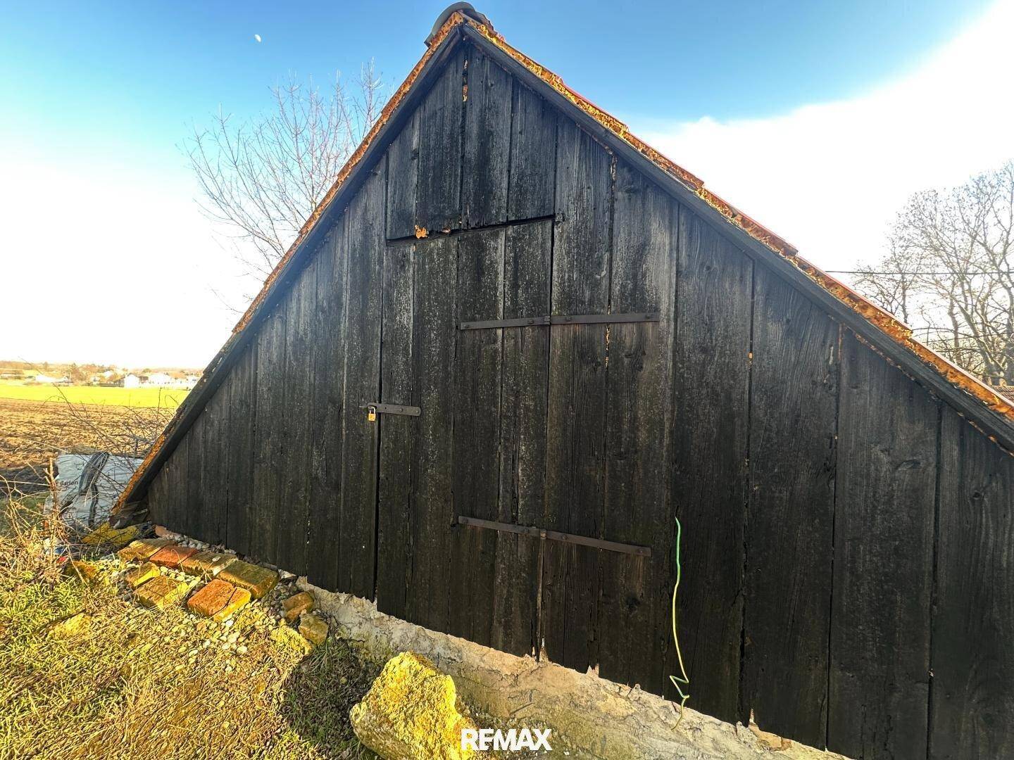 Altes, dunkles Holzgebäude mit Satteldach und rustikalen Holztüren, umgeben von Natur.