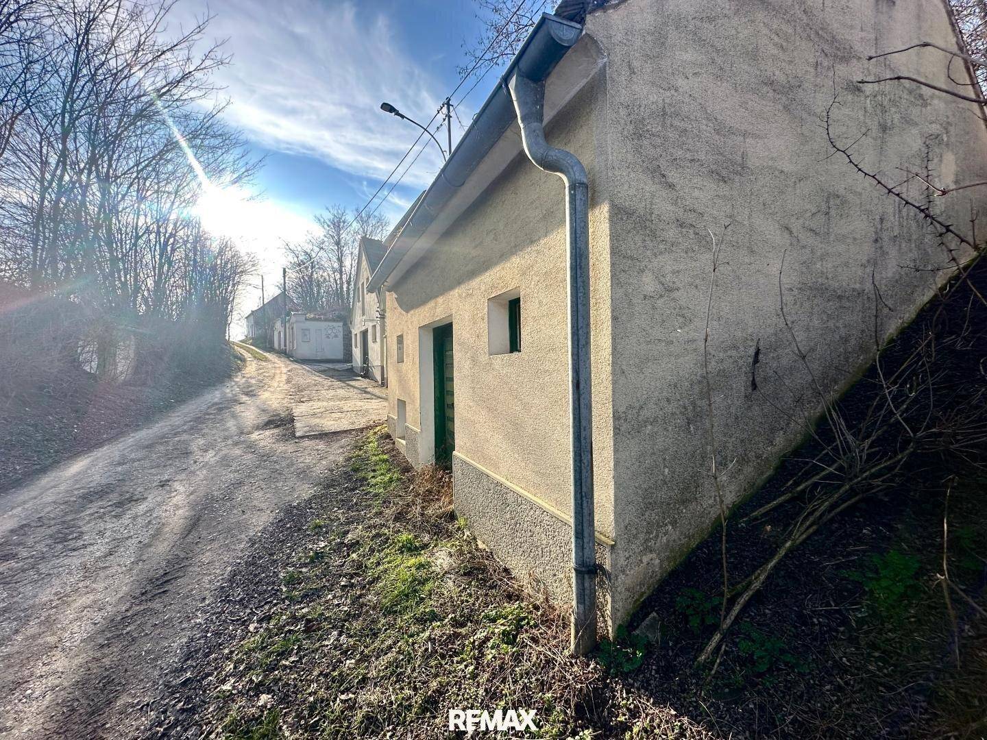 Schmaler Feldweg führt an einem einfachen Gebäude mit Putzfassade und Regenrinne vorbei.