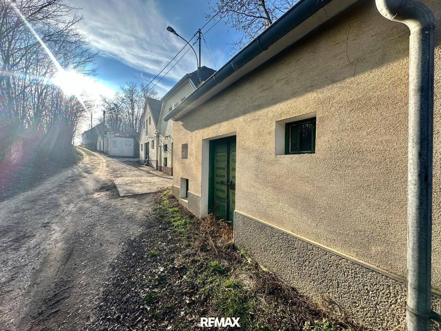 Einfaches Gebäude mit beiger Putzfassade und grüner Tür an einem sonnigen Feldweg.