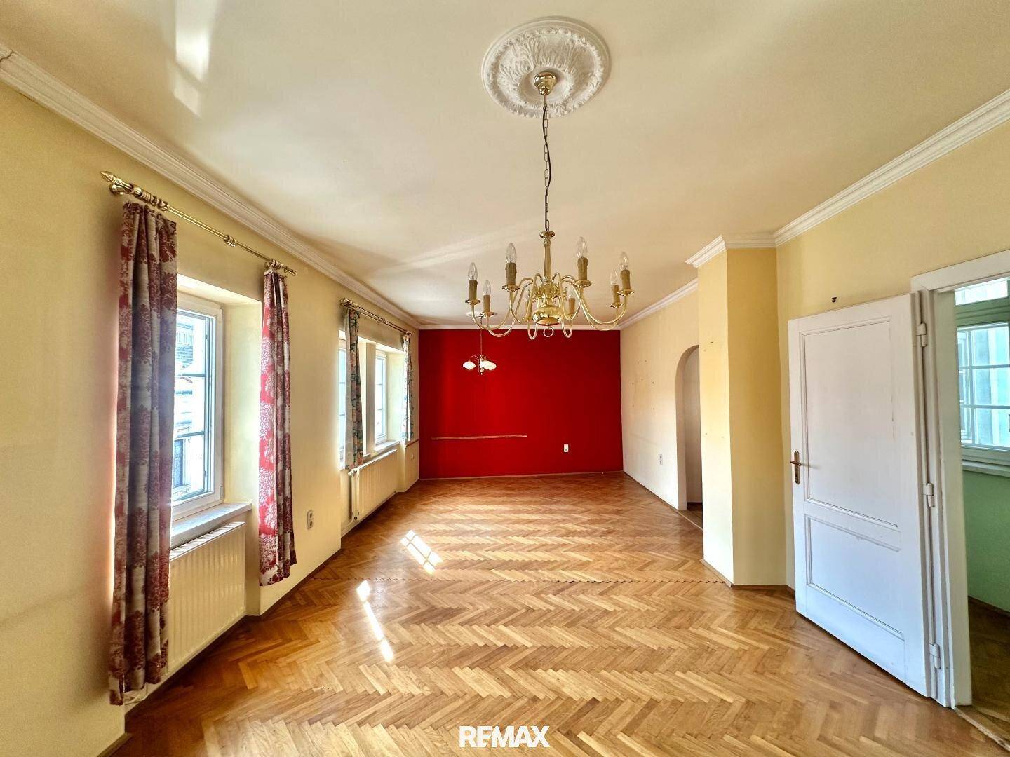 Geräumiges Wohnzimmer mit Fischgrätparkett, roter Akzentwand und hohen Decken, bereit zur Gestaltung.