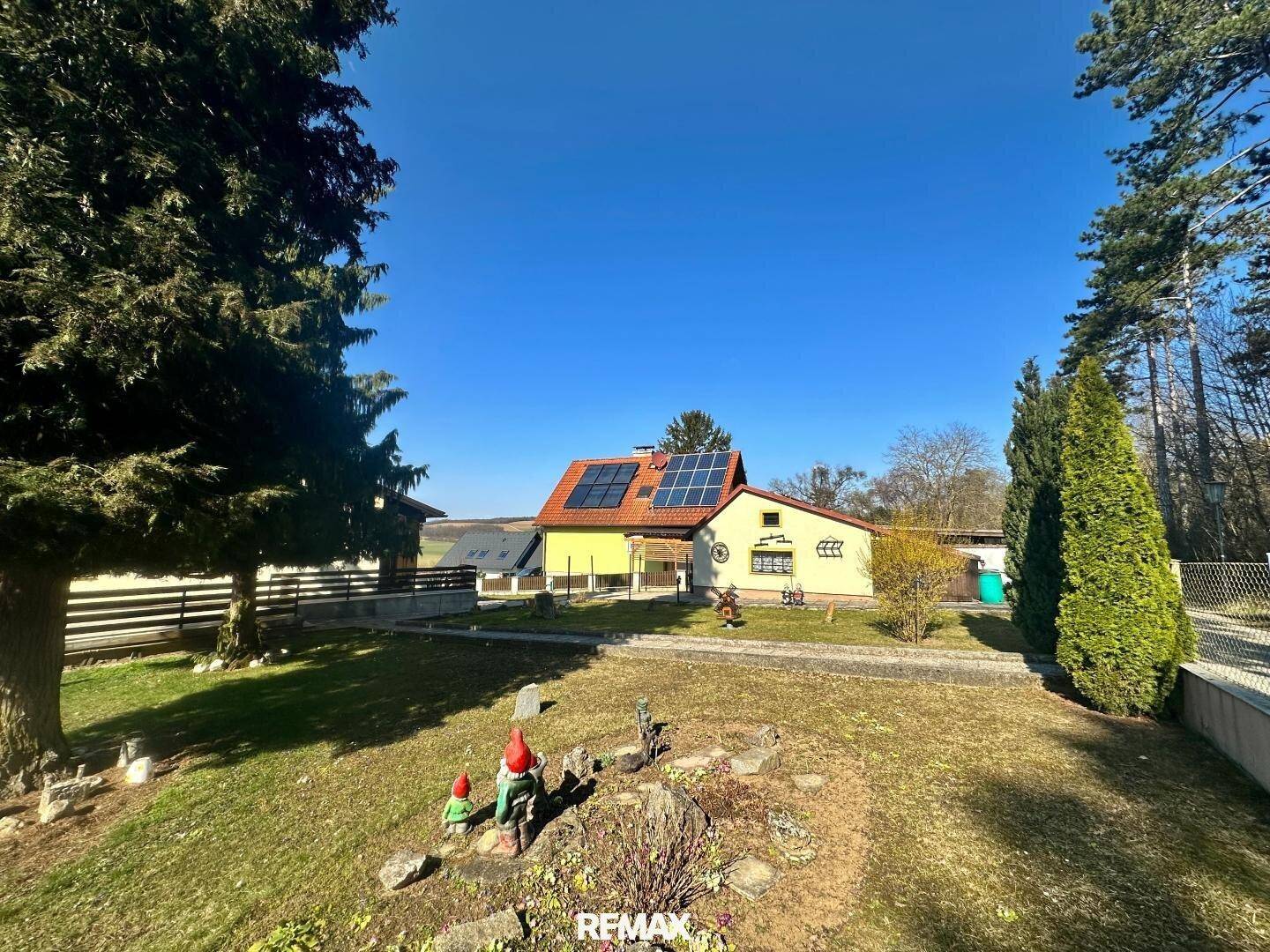 Haus mit gelber Fassade, Satteldach und Solaranlage, umgeben von einem grünen Garten mit Bäumen.