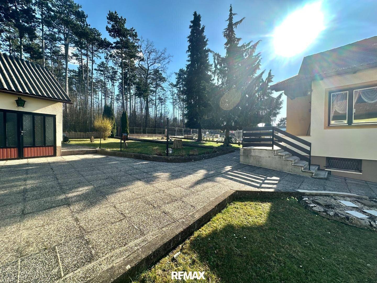 Hausansicht mit großer gepflasterter Fläche, umgeben von Rasen und Bäumen an einem sonnigen Tag.
