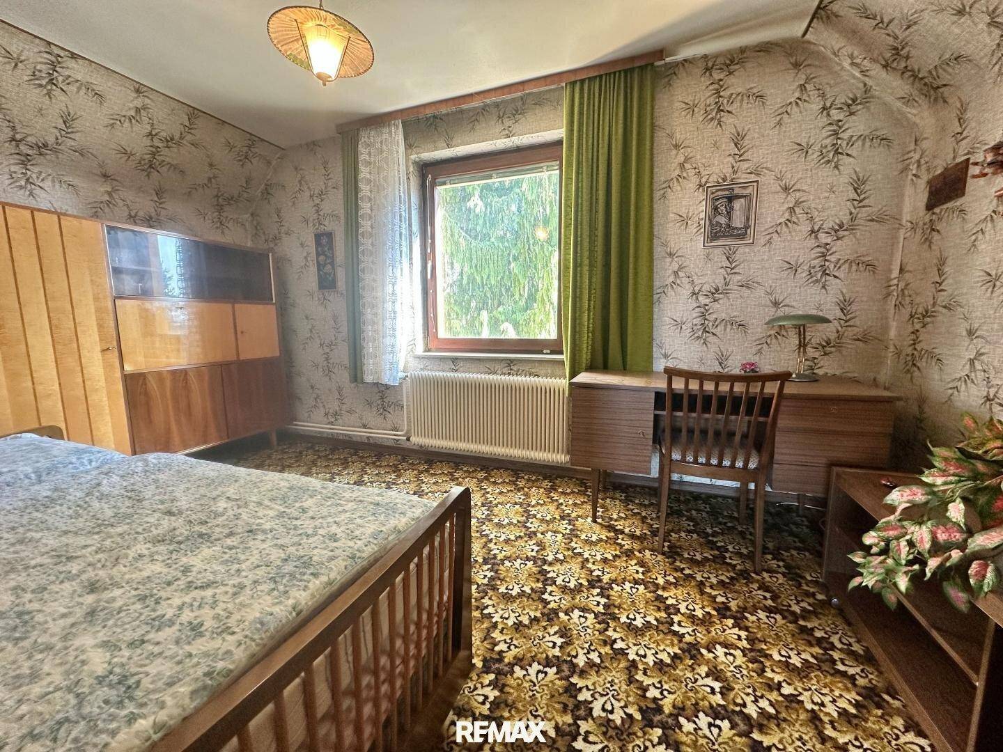 Schlafzimmer mit gemustertem Teppich, Holzbett, Schreibtisch und grünen Vorhängen.