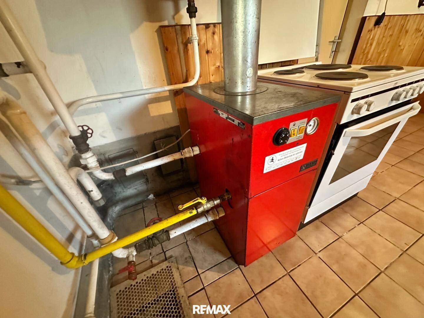 Heizungsraum mit rotem Boiler, sichtbaren Rohren und einem alten Elektroherd auf Fliesenboden.