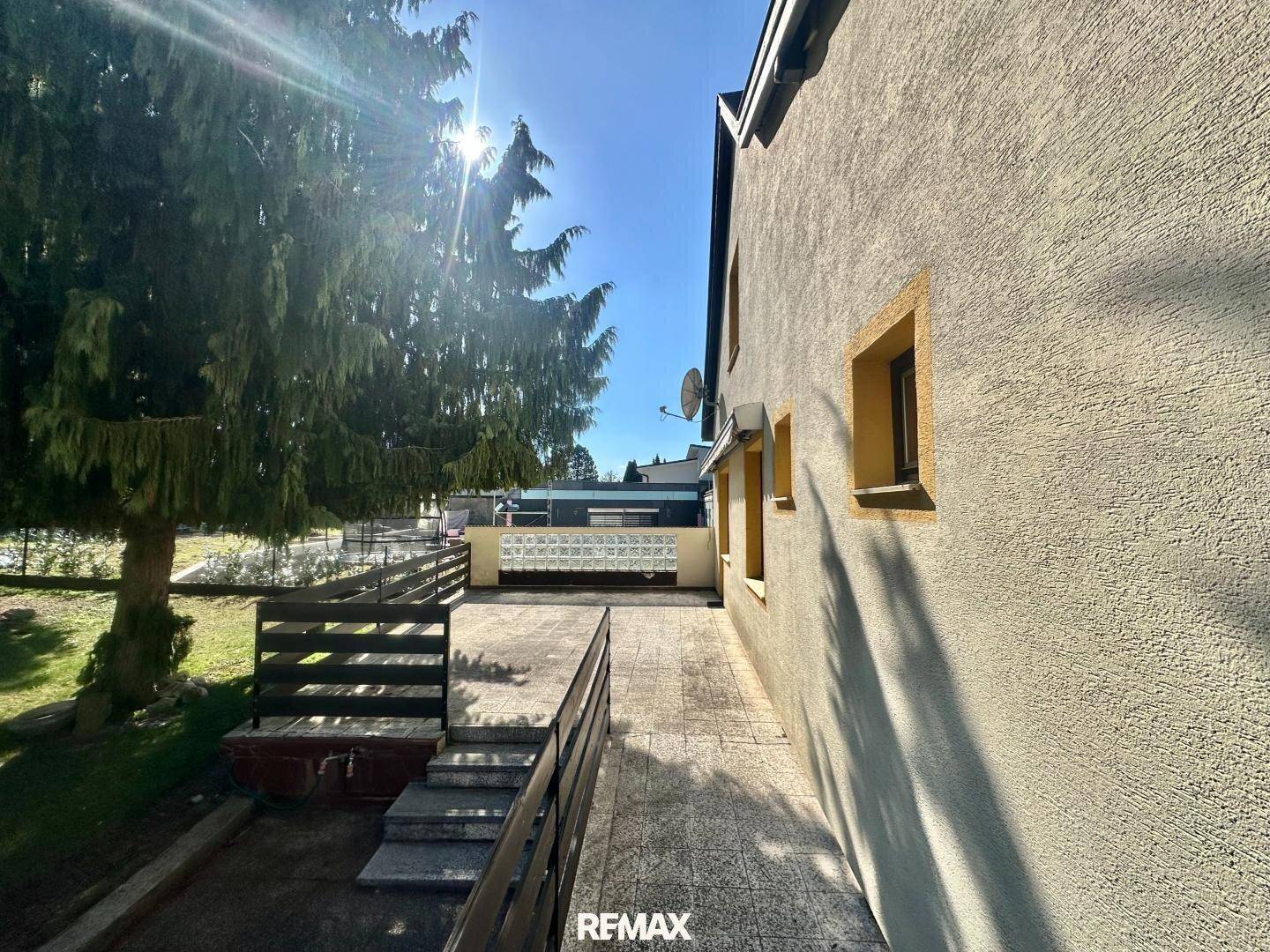 Sonnige Außenterrasse mit gepflastertem Weg, Holzgeländer und Blick auf den grünen Garten.