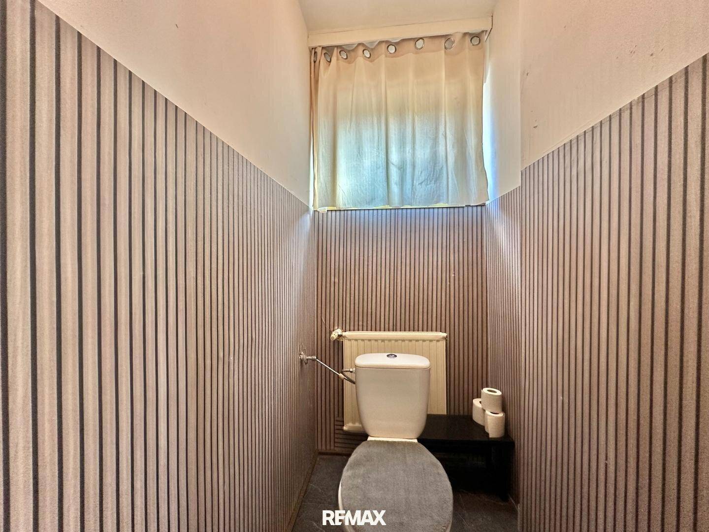 Kleines WC mit Toilette, Heizkörper und einem Fenster, die Wände sind gestreift tapeziert.