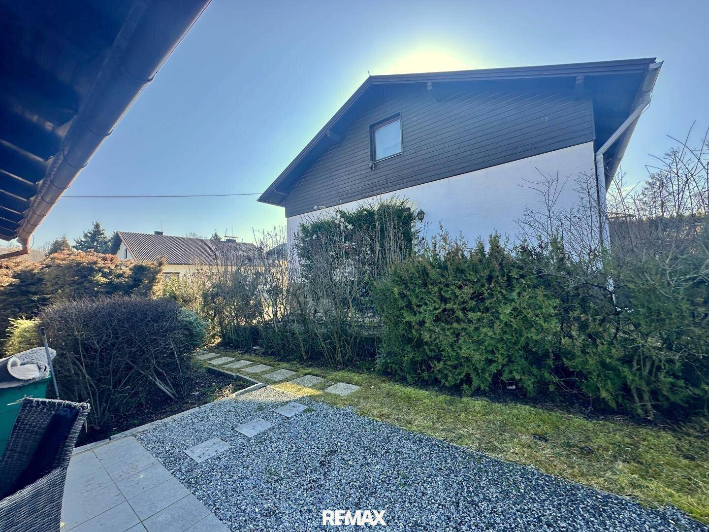 Seitenansicht des Hauses mit gepflastertem Bereich und grünen Hecken unter strahlendem Himmel.