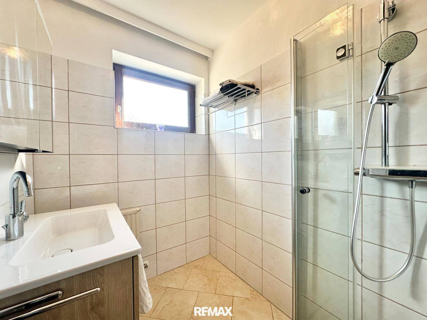 Badezimmer mit Einzelwaschbecken, Duschkabine und einem Fenster, hell gefliest.