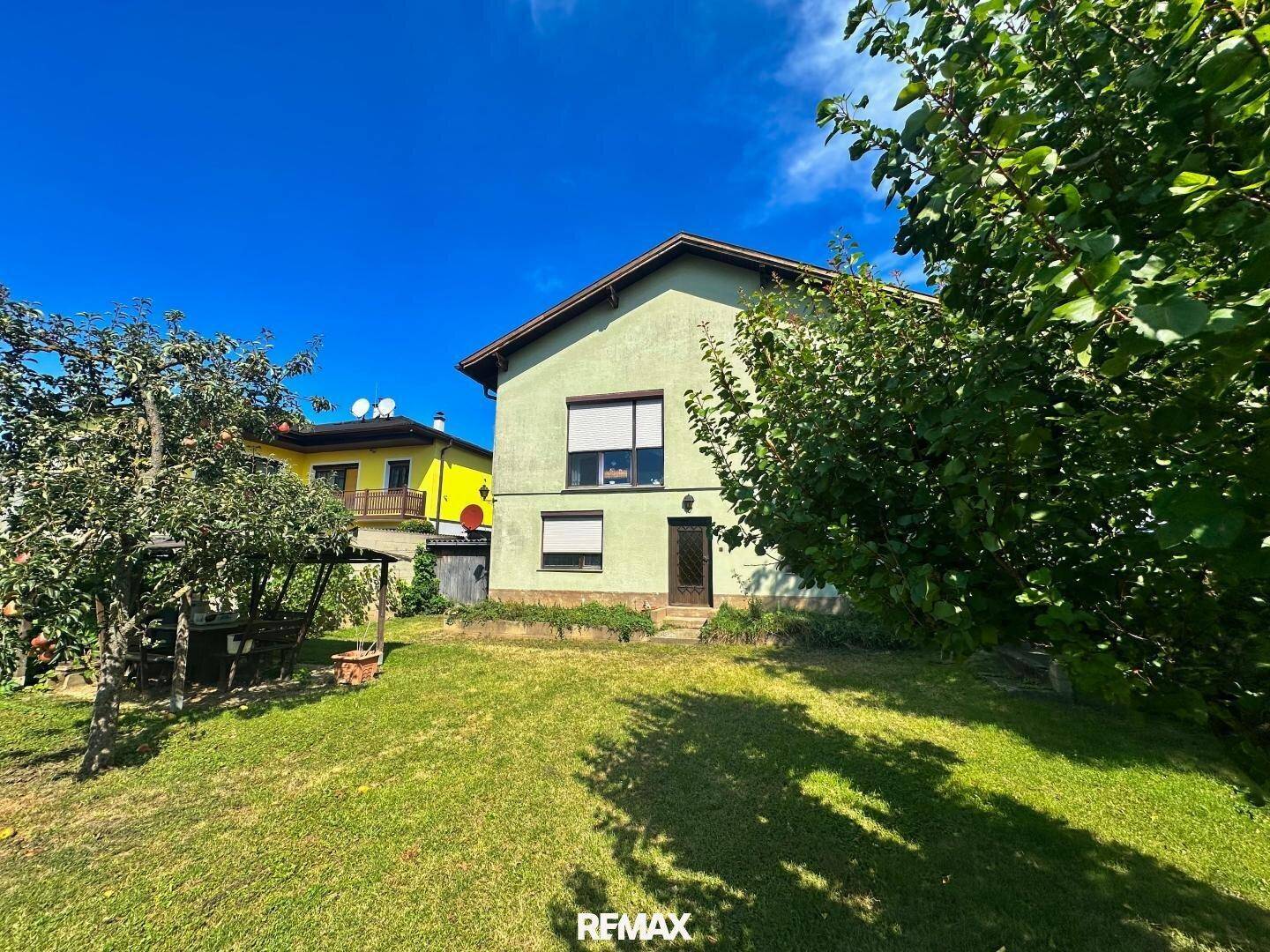 Haus mit großem Garten, umgeben von Bäumen und einer weiten Rasenfläche unter blauem Himmel.