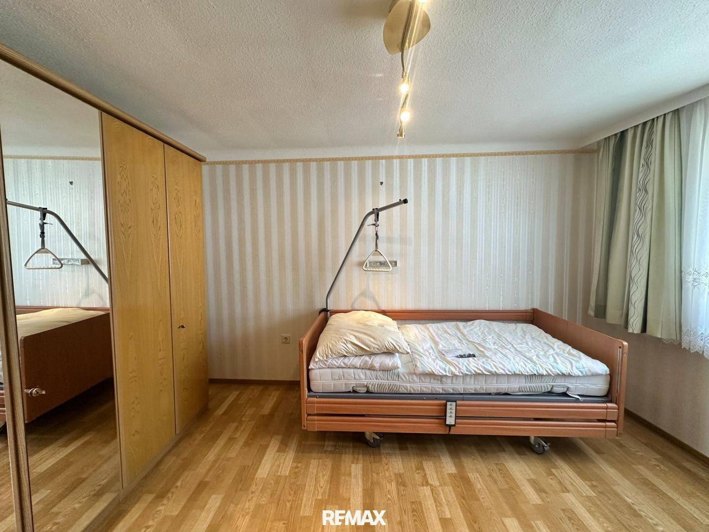 Schlafzimmer mit Pflegebett und großem Holzschrank, ausgestattet mit gestreifter Tapete.