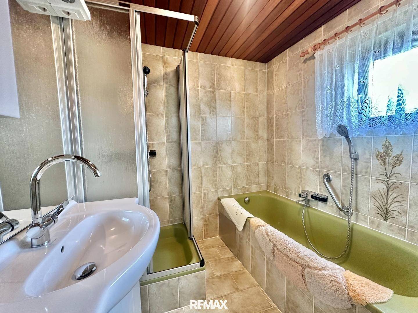 Badezimmer mit grüner Badewanne, Duschkabine und Waschbecken, umgeben von beigen Fliesen und Holzdecke.