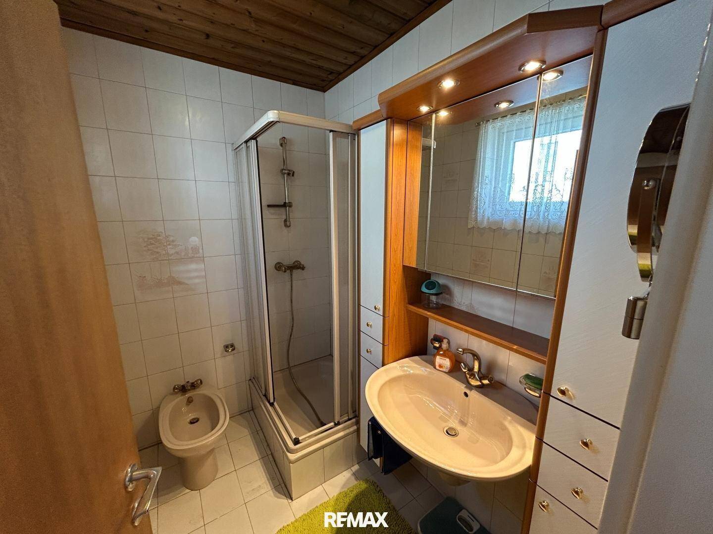 Gefliestes Badezimmer mit Duschkabine, Waschbecken, Spiegelschrank und Bidet, mit Fenster.
