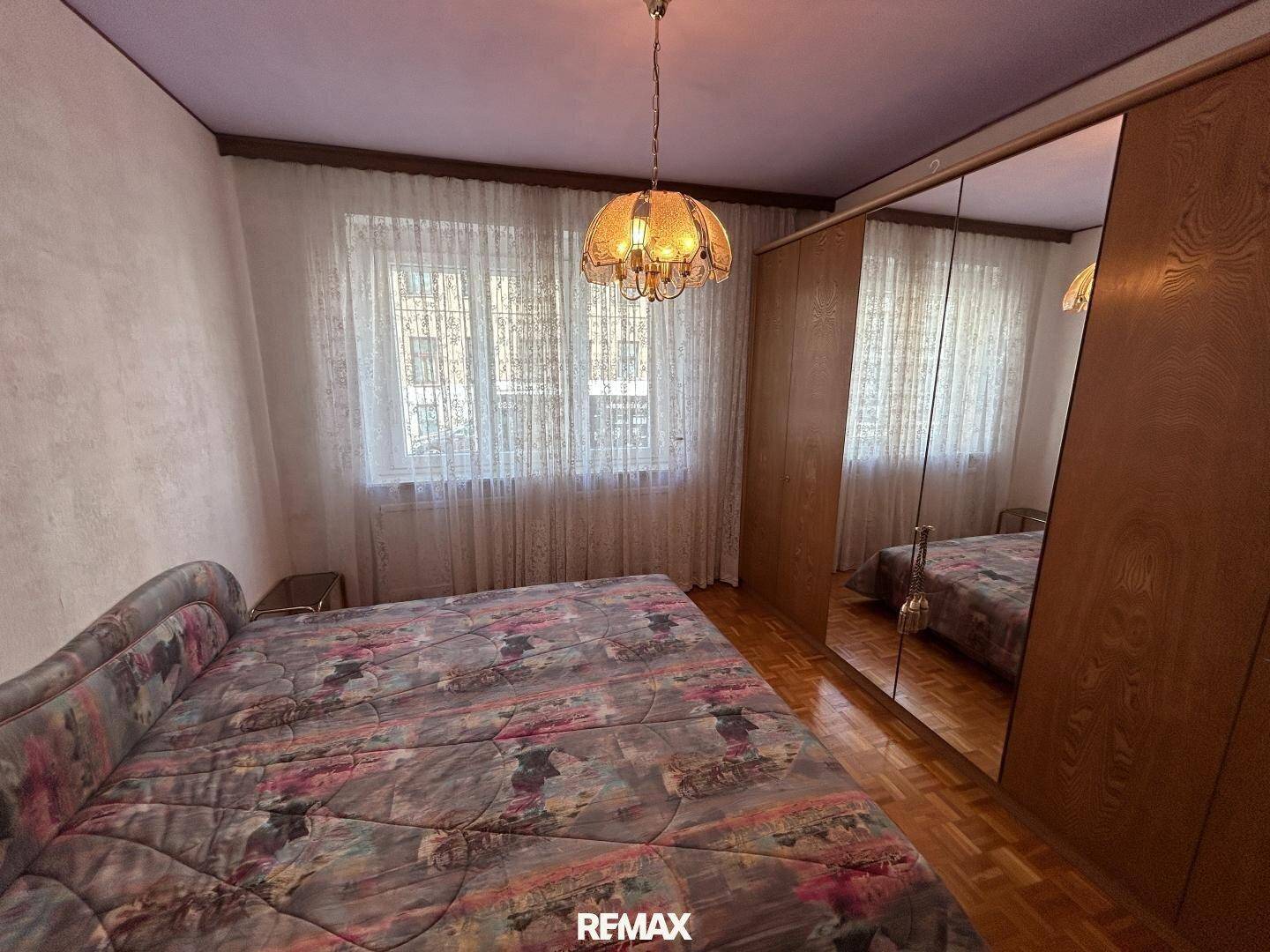 Schlafzimmer mit Doppelbett, großem Spiegelschrank und Parkettboden, hell durch das Fenster.