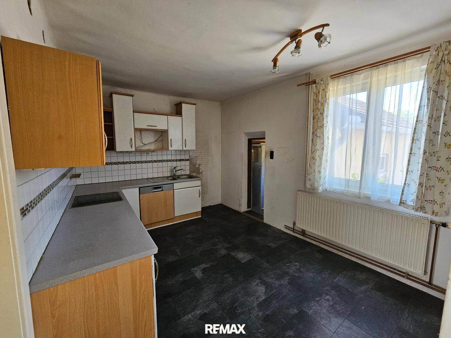 Küche mit älteren Holzschränken, Fliesenboden und Fenster mit Vorhängen.