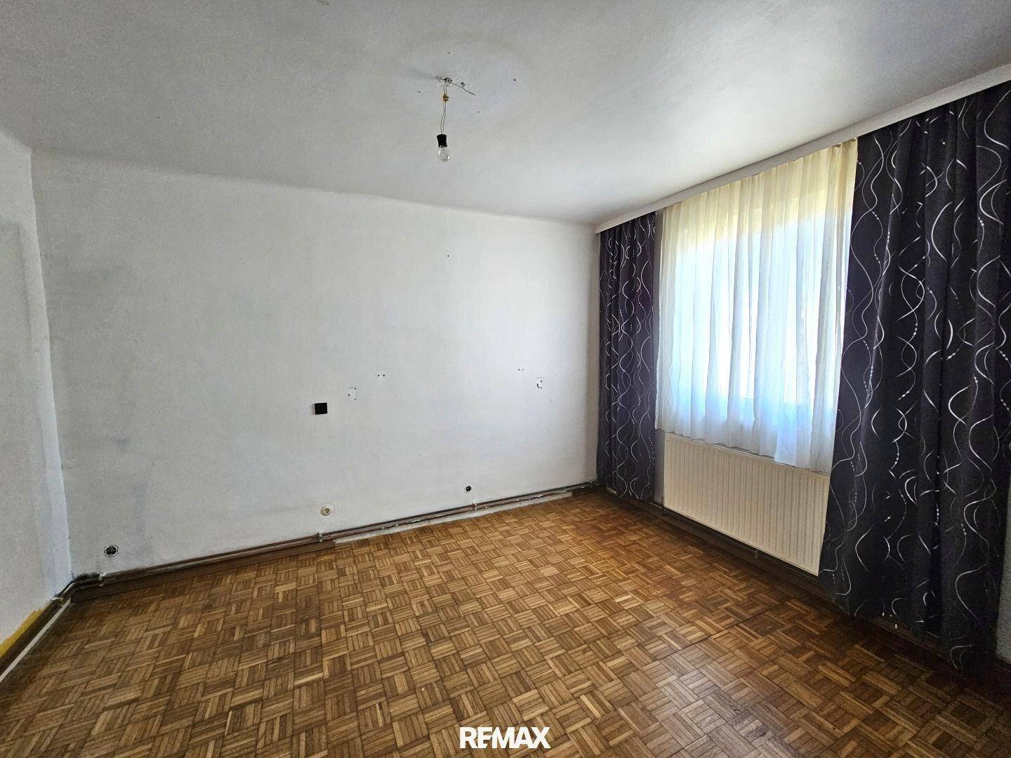 Schlafzimmer mit Parkettboden, großem Fenster und dunklen Vorhängen.