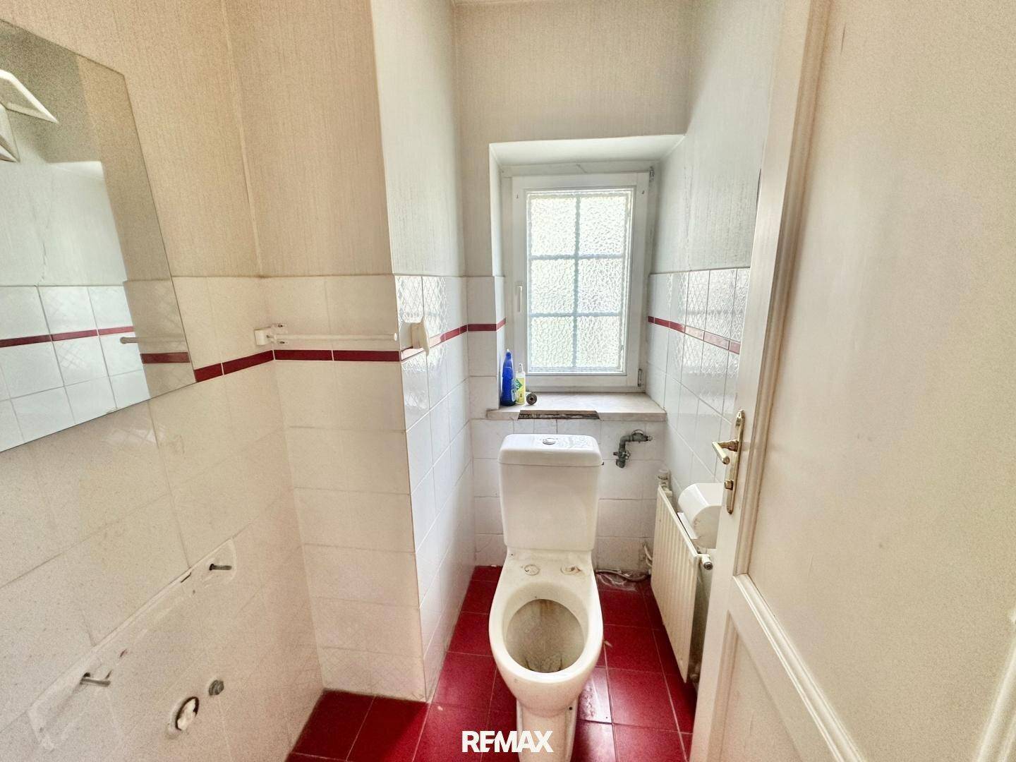 Separate Toilette mit roten Bodenfliesen, Fenster und einfacher Ausstattung.