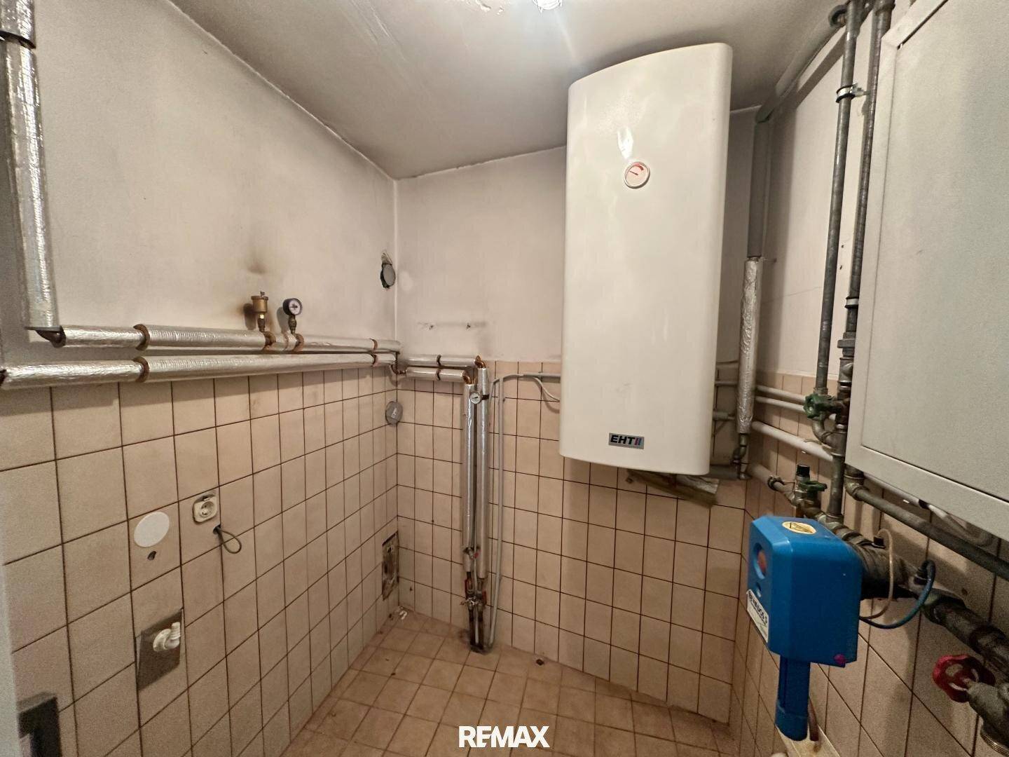 Technikraum mit Warmwasserbereiter, sichtbaren Rohren und gefliesten Wänden.