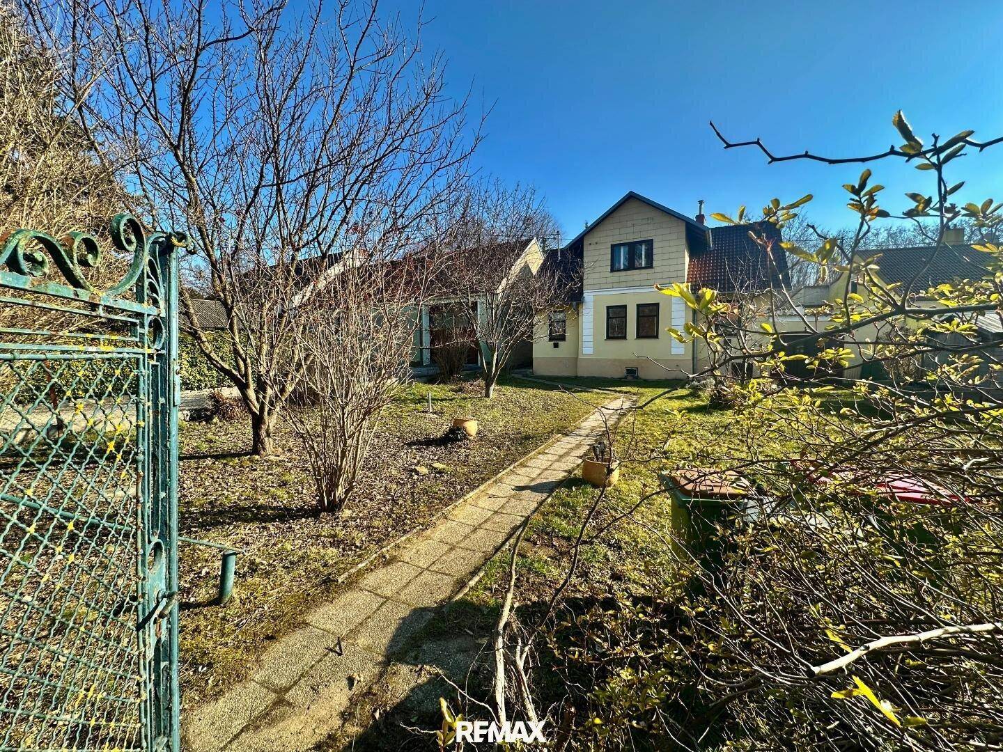 Gartenweg führt zum Haus, umgeben von Bäumen und Rasenflächen unter strahlend blauem Himmel.