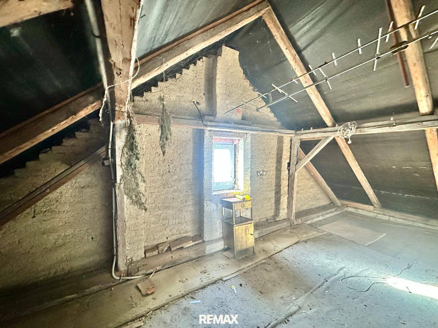 Unausgebauter Dachboden mit sichtbaren Holzbalken und einem kleinen Fenster, bietet Ausbaupotenzial.