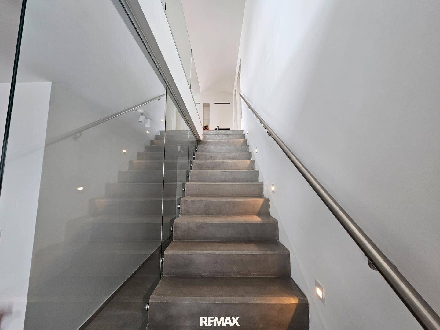 Moderne Treppe mit Betonstufen und Glasgeländer, beleuchtet von integrierten Wandleuchten.