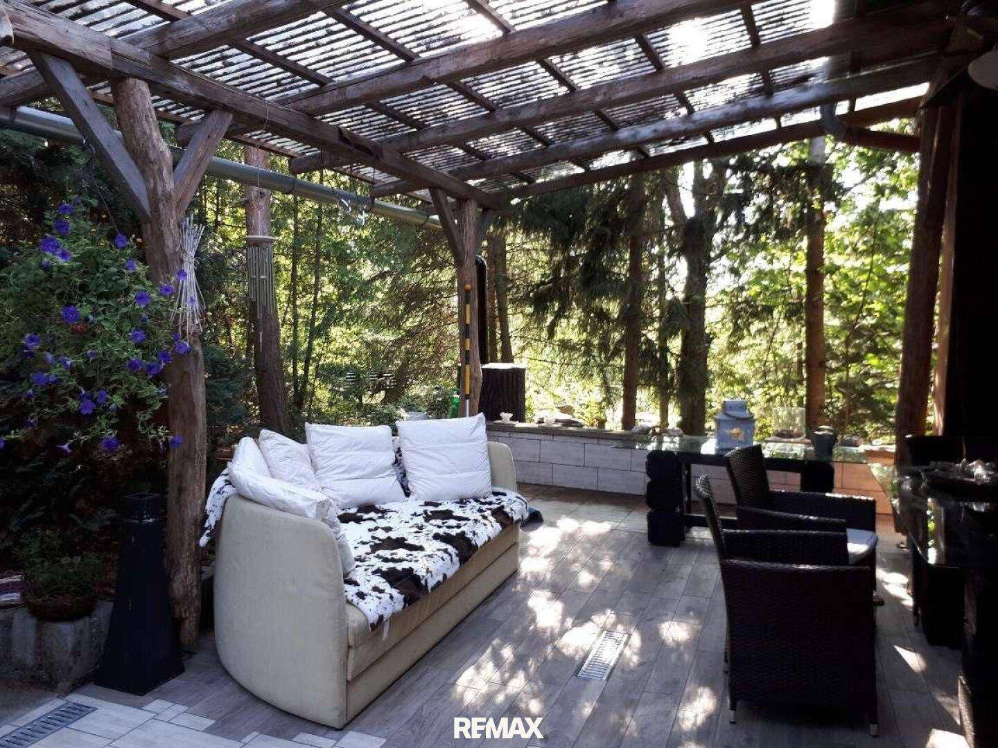 Überdachte Terrasse mit bequemen Möbeln, umgeben von Bäumen und viel Grün.