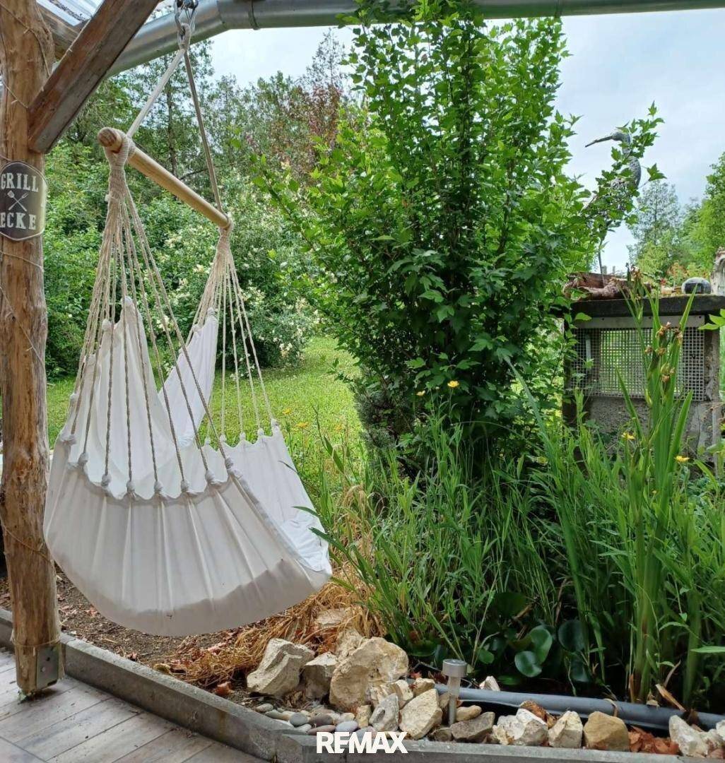 Hängesessel im Garten unter einer Pergola, umgeben von grünen Pflanzen und Felsen.