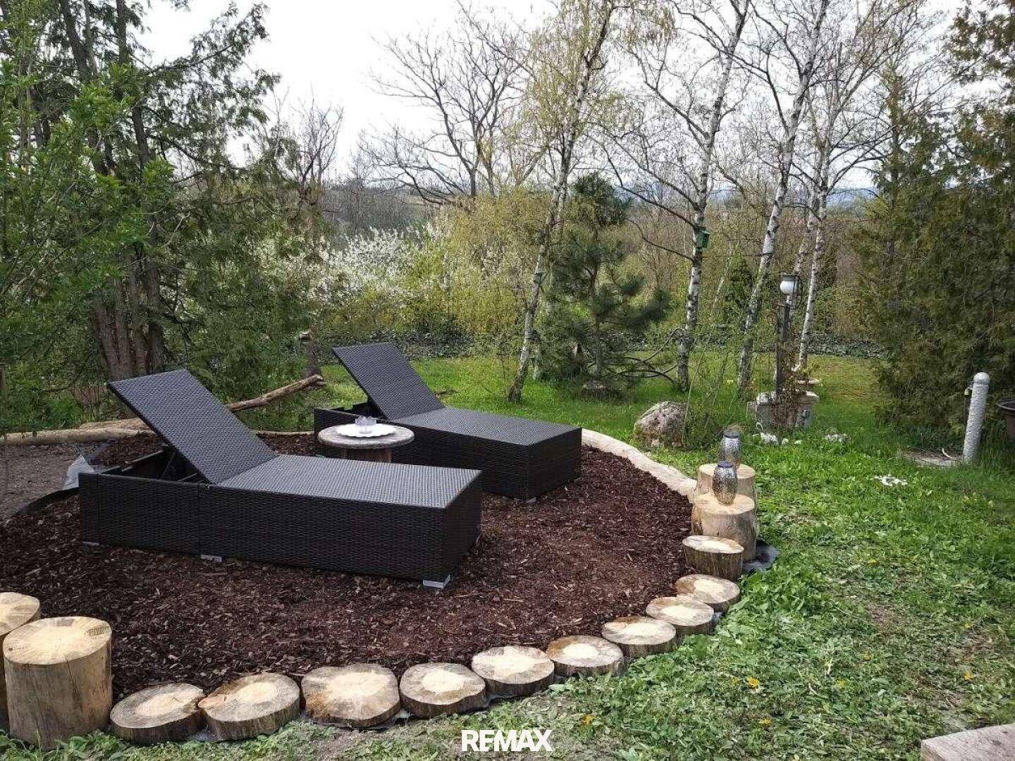 Entspannungsbereich im Garten mit Liegestühlen auf Rindenmulch und Baumstamm-Dekoration.